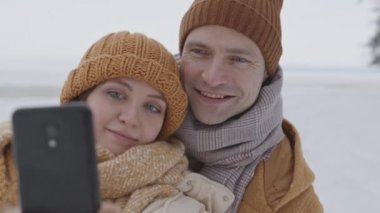 Medium closeup with slowmo of young happy Caucasian couple taking winter selfie portrait on smartphone outdoors on cold day