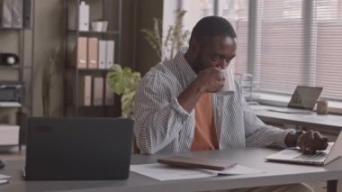 Orta yavaş çekimde Afro-Amerikan iş adamı kahve içerken ve laptopta mesaj atarken modern ofiste çalışırken.