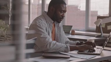 Afro-Amerikalı iş adamının günlük kıyafetleriyle finansal belgelerle çalışırken, dizüstü bilgisayarında yazarken, modern ofiste çalışma masasında otururken orta yavaş çekimleri.