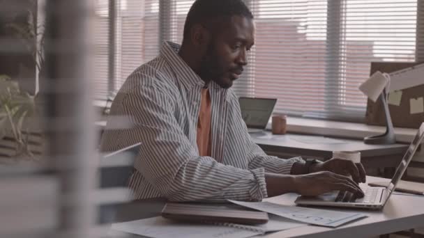 穿着便服 带着财务文件和在笔记本电脑上打字的非洲裔美国商人坐在现代办公室的办公桌前 拍了一张中等速度的照片 — 图库视频影像