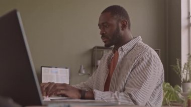 Çağdaş ofiste laptopta çalışan odaklanmış sakallı siyahi adamın orta boy yavaş çekimi.