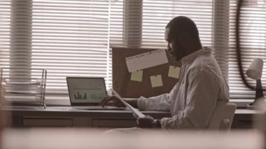 Afro-Amerikalı iş adamının dizüstü bilgisayarında finansal çizelgeler ve şemalarla çalışırken, ofisteki pencerenin önünde otururken orta yavaş çekimleri.