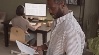 Orta yavaş çizgili tişörtlü gülümseyen Afro-Amerikalı adamın resmi. İş evrakları elinde. Modern ofiste arka planda bilgisayarla çalışan bir meslektaşla kameraya bakıyor.