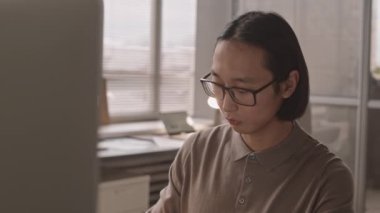 Orta boy, gözlüklü genç Asyalı adamın modern ofiste çalışan bilgisayar monitörüne bakıp arka planda oturan Afro-Amerikan meslektaşıyla yakın çekim.