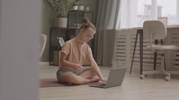 11岁女孩通过笔记本电脑在线上瑜伽课 在舒适的家中舒适地坐在瑜伽垫上摆出莲花姿势的慢镜头 — 图库视频影像