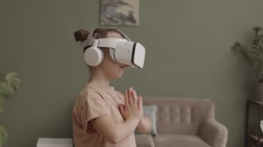 VR kulaklık takmış, kulaklık takmış, dua eden ellerle evde meditasyon yapan küçük bir kızın orta yavaş çekimi.