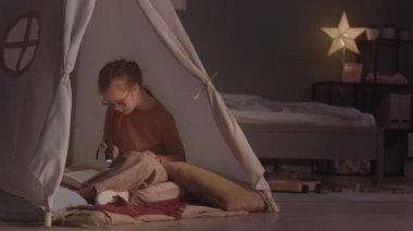 Geceleri rahat bir yatak odasında çadırda otururken elinde meşaleyle kitap okuyan güzel bir kızın yavaş çekimleri.