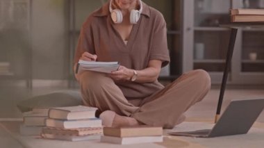 Rahat rahat giyinen yetişkin bir kadının elinde kitap yığınları ve notlarla dizüstü bilgisayarında tek başına çalışırken çekilmiş yavaş çekimleri.