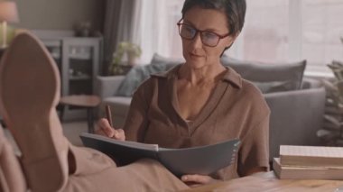 Yetişkin, beyaz bir kadının laptopta eğitim videosu izlerken not aldığı, evden ders aldığı, bacakları masanın üzerinde rahat bir şekilde oturduğu,