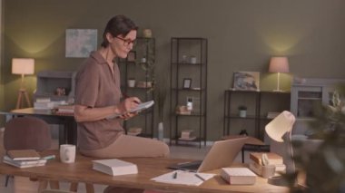 Gözlüklü orta yaşlı bir kadının laptopunda eğitim videosu izlerken not alırken, evden internetten ders çalışırken orta boy bir fotoğrafı.