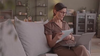 Odaklanmış olgun bir kadının laptopunda eğitim videosu izlerken not aldığı, rahat oturma odasındaki kanepede oturduğu orta derece yavaş bir fotoğraf.