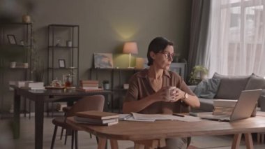 Elinde bir fincan sıcak çayla masa başında oturmuş, üzerinde bir yığın belge ve dizüstü bilgisayarla rahat bir evde tek başına ders çalışan olgun, beyaz bir kadının orta yavaş çekimleri.