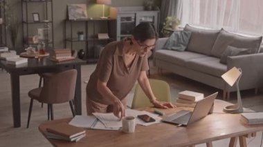 Kafkasyalı orta yaşlı bir kadının elinde notlar ve eğitim materyalleriyle evde ders çalışırken dizüstü bilgisayardan eğitim videosu izlediği orta yavaş bir fotoğraf.