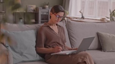 Orta yaşlı beyaz kadın laptopta çalışırken ve rahat oturma odasındaki kanepede otururken notlar alırken.