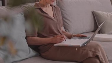 Yetişkin bir kadının internet dersinde not aldığı, rahat bir evde kanepede oturduğu yavaş çekimde çekilmiş.