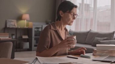 Orta yaşlı bir kadının laptopta video dersi izlerken çay içerken, çalışma materyalleri yığını ile masada otururken, evden tek başına ders çalışırken orta yavaş çekimleri.