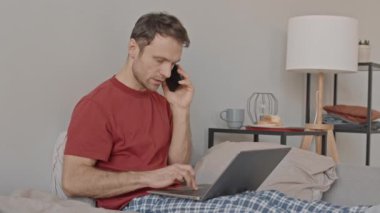 Kafkasyalı iş adamının kareli pijama pantolonlu, akıllı telefonlu, dizüstü bilgisayarlı, yatakta sabahları otururken görüntüsü.