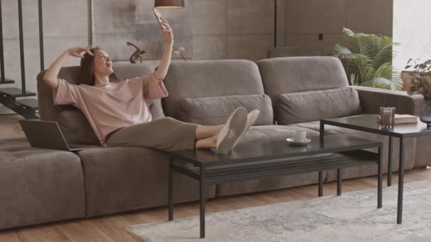 身着便衣的白人年轻女子在家工作时 在智能手机上用有趣的自拍照 坐在沙发上 还带着笔记本电脑坐在现代公寓里 拍下了一张速度慢的照片 — 图库视频影像