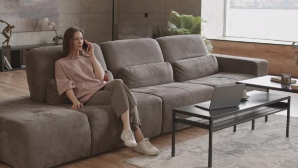 在现代公寓的沙发上用手机交谈的年轻白人妇女的慢镜头 — 图库视频影像