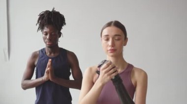 Orta yavaş çekimde, yüksek teknolojili protez kollu beyaz bir kadın Afro-Amerikalı erkek arkadaşıyla birlikte meditasyon yaparken, beyaz arka planda dua eden elleri olan,