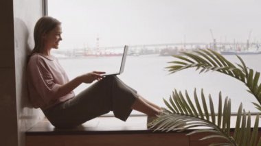 Yan görüntüde gülümseyen genç kadın videosu dizüstü bilgisayarla konferans yaparken panoramik pencerenin yanında deniz manzaralı.