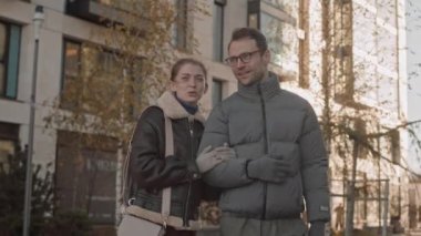 Güneşli bir sonbahar gününde dışarıda yürürken mutlu, genç, beyaz bir çiftin sohbet ederken orta yavaş çekimleri.