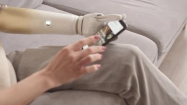 Biyonik kollu, akıllı telefonunda yemek dağıtım uygulaması kullanan beyaz bir kadının evdeki kanepede otururken yavaş çekimleri.