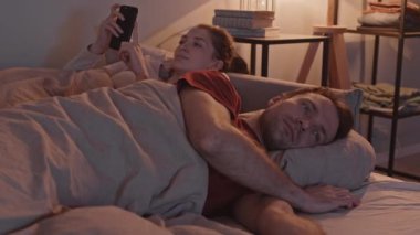 Yavaş çekimde, genç romantik çift gece yatakta birlikte yatarken, erkek televizyon izlerken kız arkadaşı ona akıllı telefonundan bir şey gösterip birlikte gülüyor.