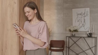 Orta yavaş çekimde, neşeli genç bir kadının arkadaşlarıyla akıllı telefondan sohbet ederken, evde otururken.