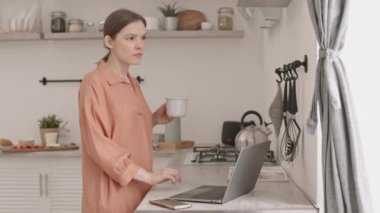 Genç, beyaz bir kadının laptopta çalışırken rahat, aydınlık bir mutfakta yemek masasında otururken orta yavaş çekimleri.