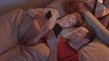 Beyaz tenli genç bir çiftin gece yatakta uzanırken akıllı telefonlar kullanıp sohbet etmelerinin en iyi görüntüsü.
