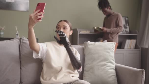 中慢镜头 年轻迷人的白种人女子 拥有仿生手臂 在智能手机上自拍 坐在沙发上 而她的非洲裔美国男友则背景看书 — 图库视频影像
