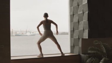 Dikiz manzaralı, sportif genç bir kadının güzel deniz manzaralı panoramik pencerenin önünde esneme egzersizleri yaptığı yavaş çekim.