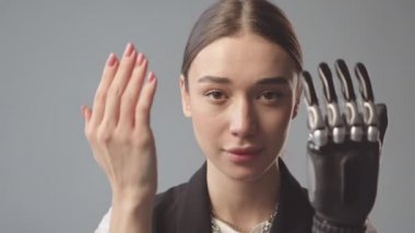 Yavaş yavaş, yüzünü ellerle kaplayan yüksek teknolojili protez kollu ve gri arka planda duran kameraya bakan güzel beyaz bir kadının portresi.