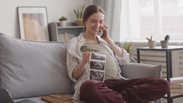 中低速ショットの笑顔白人妊娠中の女性保持超音波スキャンの彼女の赤ちゃんと話してスマートフォンで夫と 座っていますソファに居心地の良い家 — ストック動画