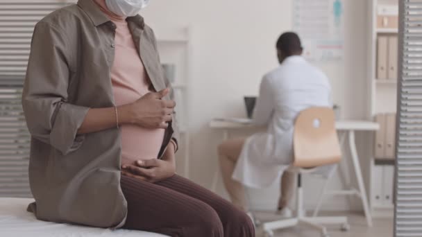 作物遅いショットの妊娠中の女性ストローク彼女の腹座っています上の医療ソファで現代的な医師のオフィス 彼女の男性婦人科医仕事で机の上に背景 — ストック動画