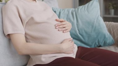 Kafkasyalı, rahat ev elbiseli hamile bir kadının kanepeye oturarak karnını okşamasının yavaş çekimleri.