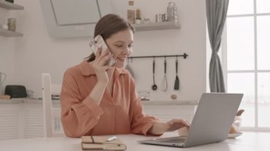 Genç iş kadınının dizüstü bilgisayarda çalışırken ve mutfak masasında otururken telefon görüşmesi yaparken orta yavaş çekimleri.
