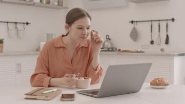 Sağlıklı bir kahvaltı yaparken laptopta çalışan beyaz bir kadının orta yavaş çekimi.