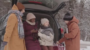 Karlı bir günde, kış ormanlarında açık araba bagajında oturup termostan sıcak çay içip sohbet eden ve birlikte vakit geçiren bir grup mutlu genç arkadaşın orta yavaş çekimi.