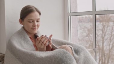 Rahat gri ekose gömlekli, pencereye bakan bir fincan sıcak çayla oturan hayalperest genç kadının orta yavaş çekimi.