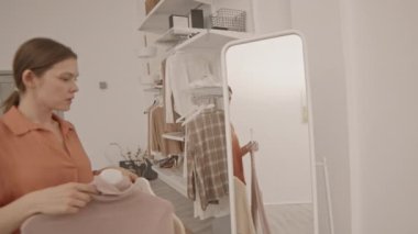 Askıları askılı genç beyaz bir kadının aynanın önünde dikilip ne giyeceğine karar verirken orta yavaş çekilen bir fotoğrafı.