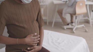 Modern doktor muayenehanesinde muayene sırasında karnını okşayan gülümseyen Afro-Amerikan kadının yavaş yavaş portresi.