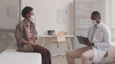 Orta yavaş çekimde, modern klinikteki randevusunda erkek jinekologla konuşurken yüzü maskeli genç Afro-Amerikalı hamile kadının karnını okşaması.