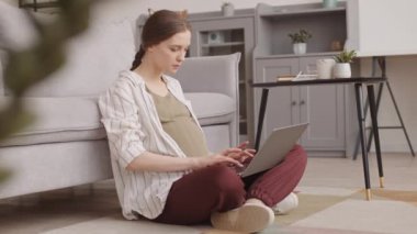 Bebek tekmelerini hissederken, gülümserken ve karnını nazikçe okşarken laptopta çalışan beyaz hamile kadının yavaş çekimleri.