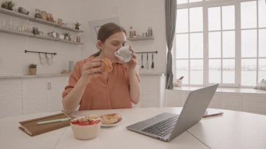 Evde kahvaltı yaparken laptoptan komik bir şey izleyen mutlu, beyaz bir kadının orta yavaş çekimi.
