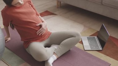Yoga minderinde dinlenen yorgun ve hamile bir kadının dizüstü bilgisayardan spor videoları izlediği yavaş çekim.