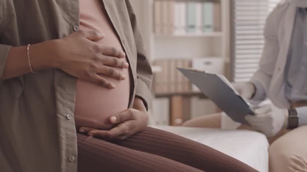 若いアフリカ系アメリカ人の妊娠中の女性の遅いショットを傾ける彼の近代的なオフィスで医師と婦人科相談中に彼女の腹を撫で — ストック動画