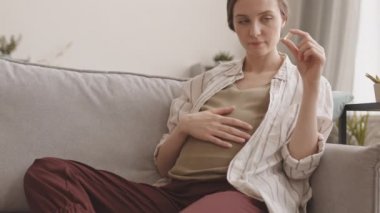 Hamile bir kadının karnını okşarken ve elinde doğum öncesi vitamin hapı tutarken yavaş çekimde evdeki koltukta otururken.