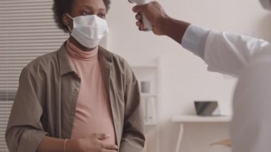 Genç Afro-Amerikalı hamile bir kadının karnını okşarken, erkek doktorun temassız kızılötesi termometreyle ateşini ölçerken,